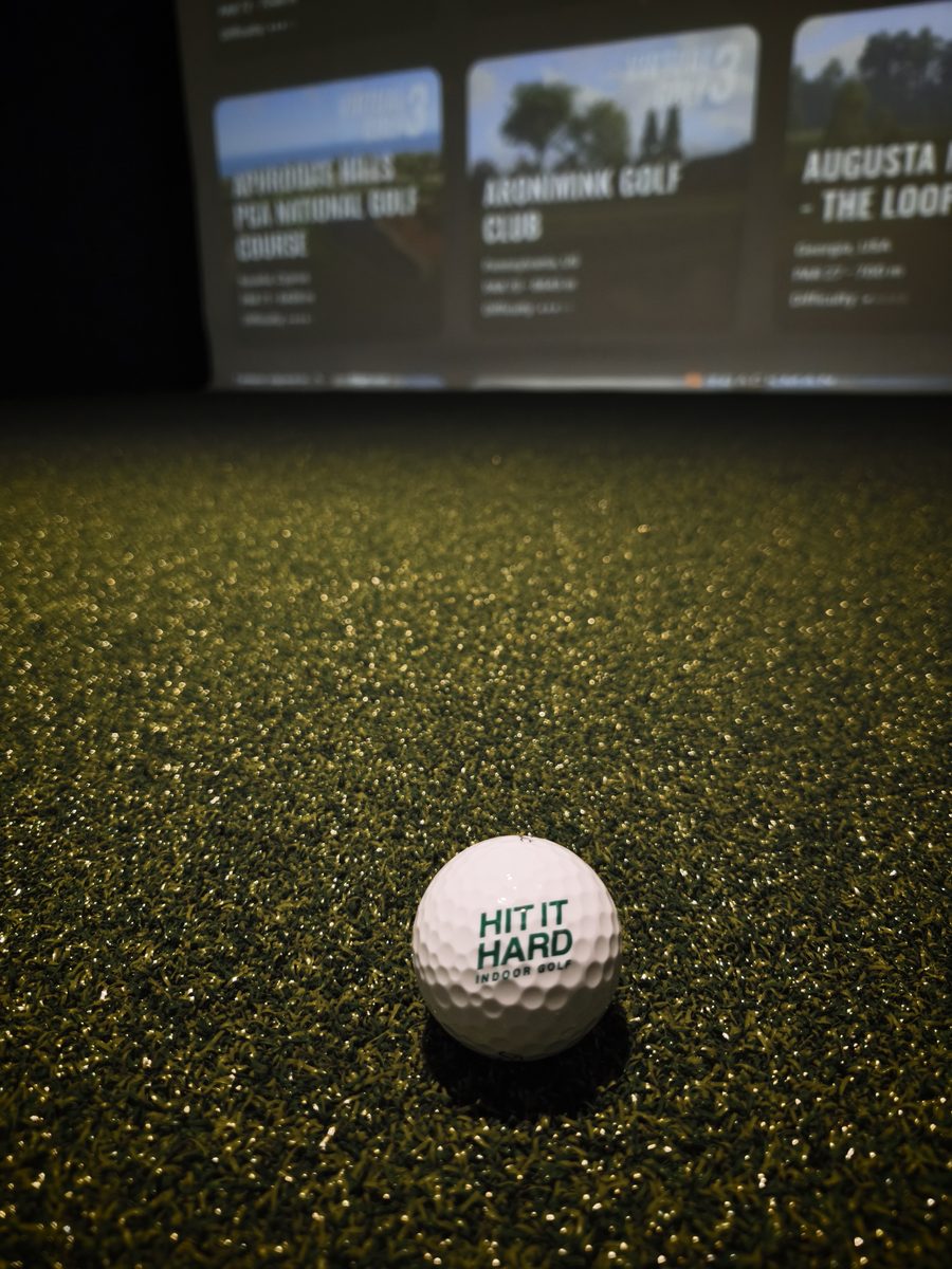 Golfball mit Hit-it-Hard-Logo auf dem Kunstrasen der Box, im Hintergrund die Platzauswahl auf der Leinwand